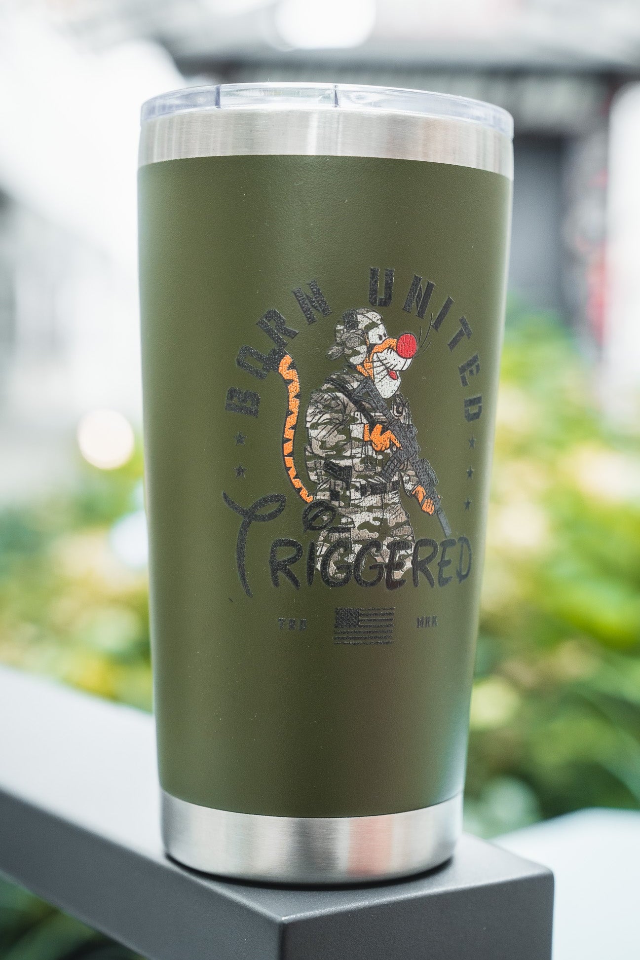 The Triggered Tumbler - 20oz by Born United Private Label is green with a metal rim, featuring a camo-clad baboon holding a rifle, "Born United Triggered" text above, an American flag below, and a blurred greenery background.