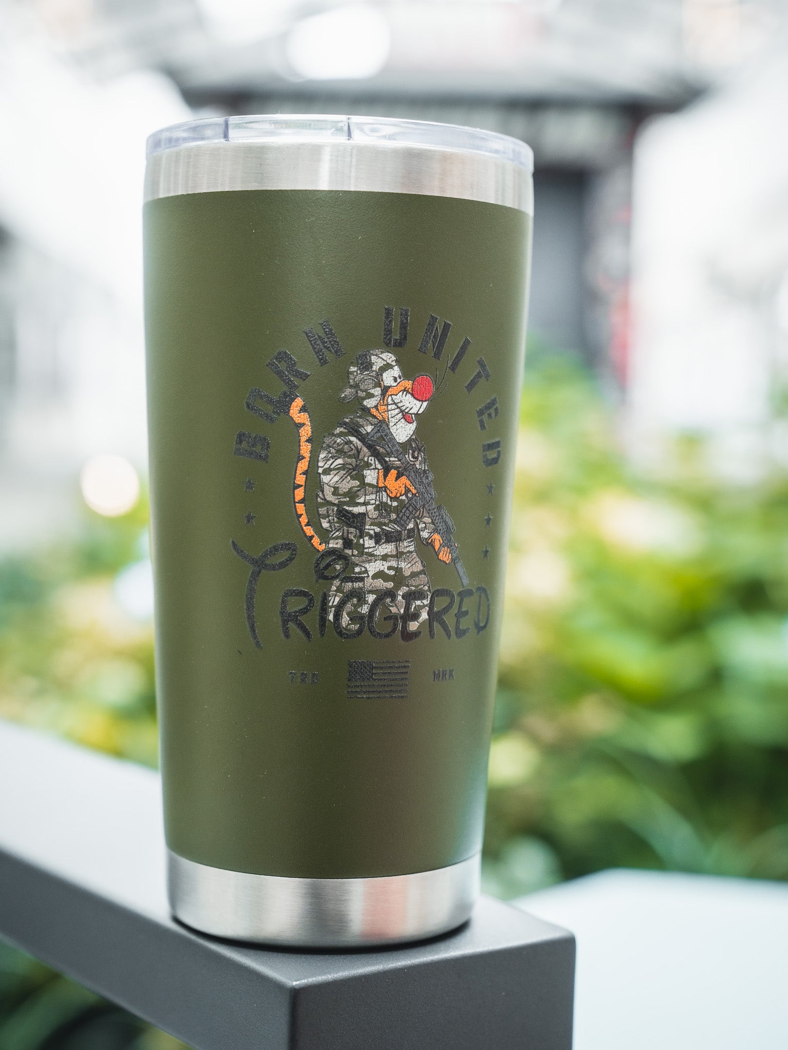 The Triggered Tumbler - 20oz by Born United Private Label is green with a metal rim, featuring a camo-clad baboon holding a rifle, "Born United Triggered" text above, an American flag below, and a blurred greenery background.