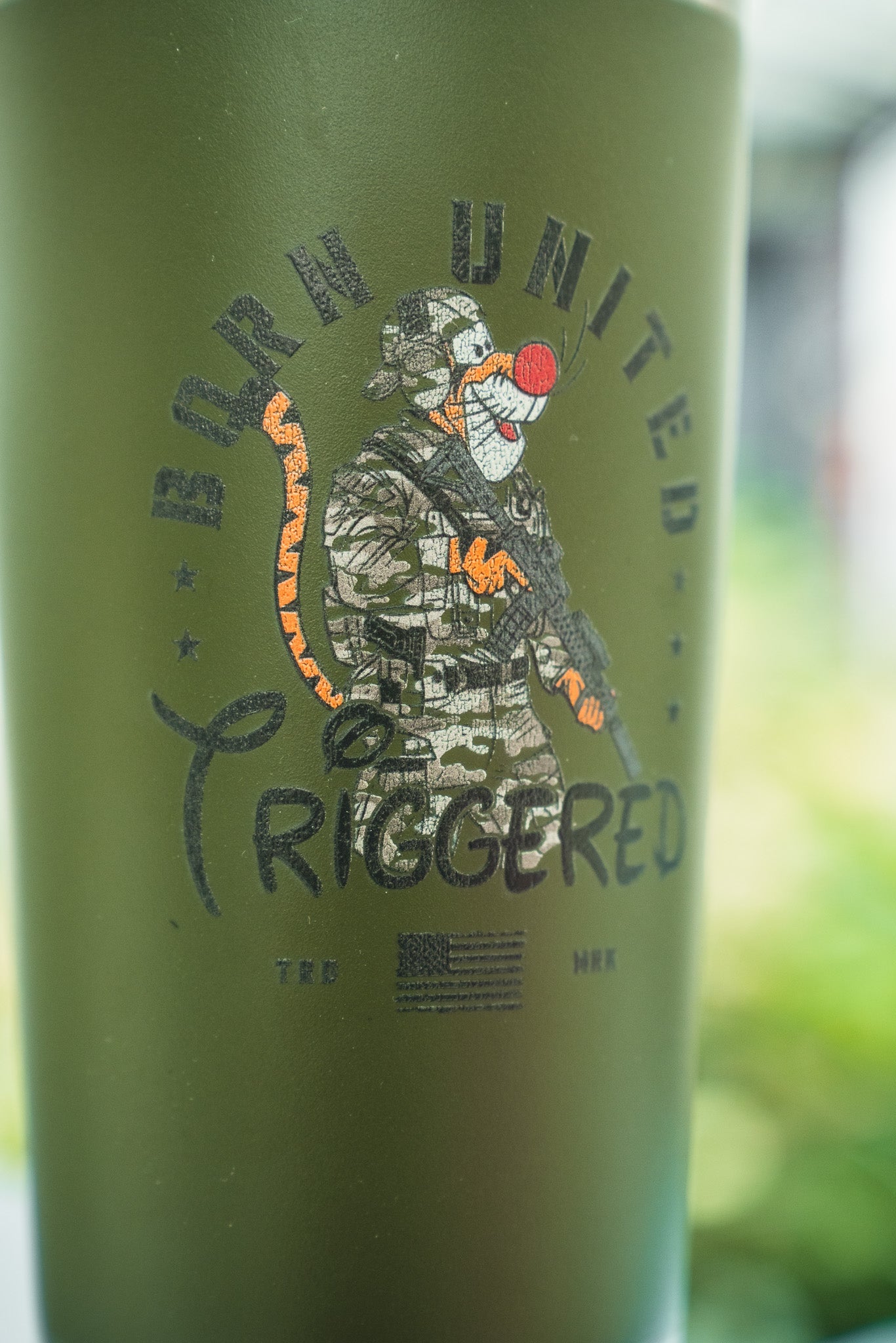 The Triggered Tumbler - 20oz by Born United Private Label features an olive green design with a tiger in military gear holding a rifle, "BORN UNITED TRIGGERED" text, and a small US flag below.