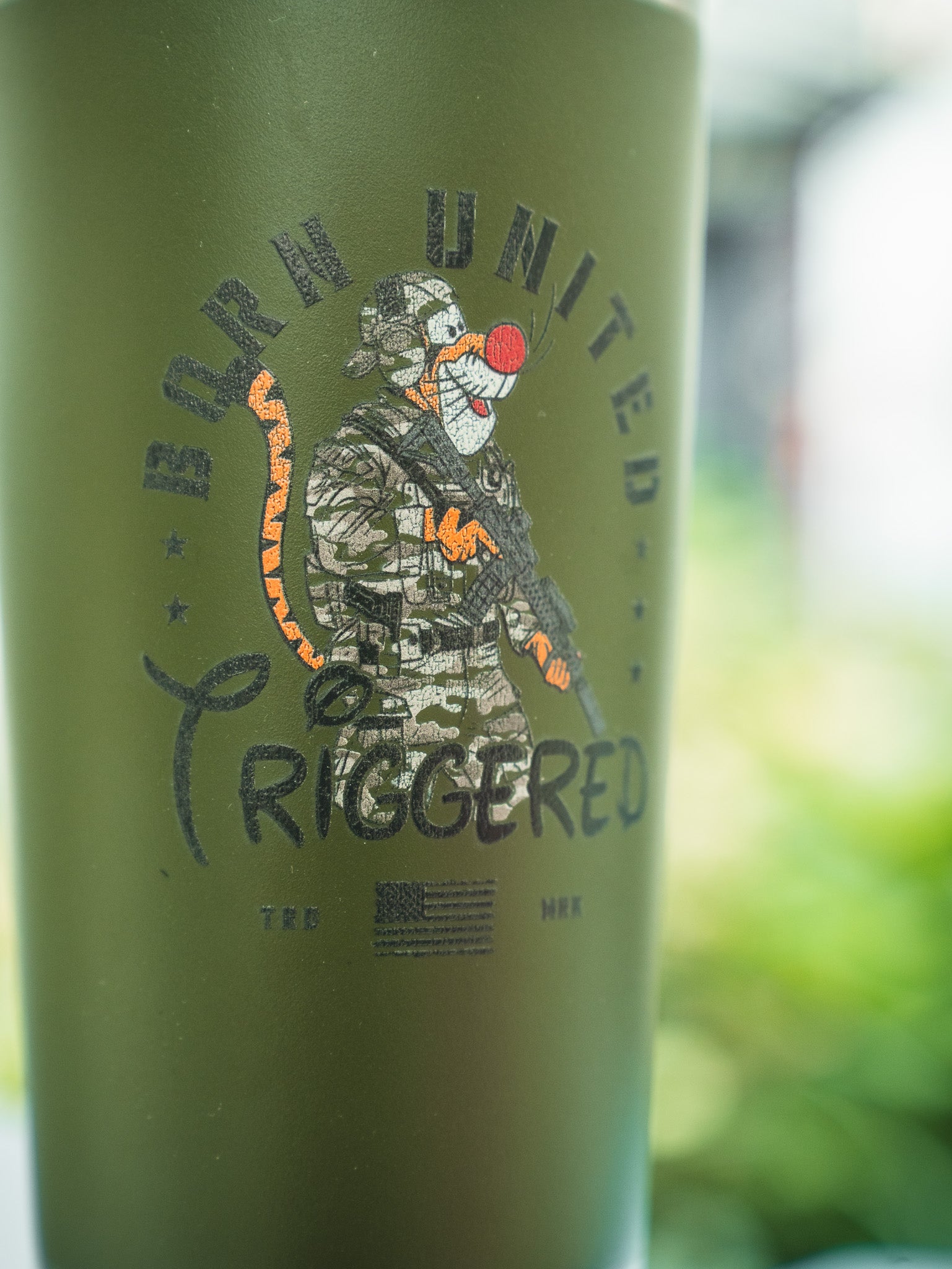 The Triggered Tumbler - 20oz by Born United Private Label features an olive green design with a tiger in military gear holding a rifle, "BORN UNITED TRIGGERED" text, and a small US flag below.