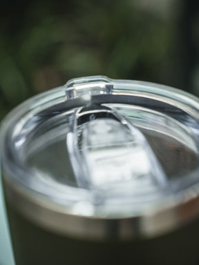 A clear plastic sliding lid tops the stainless steel 20oz Triggered Tumbler by Born United Private Label, shown against a soft green backdrop.