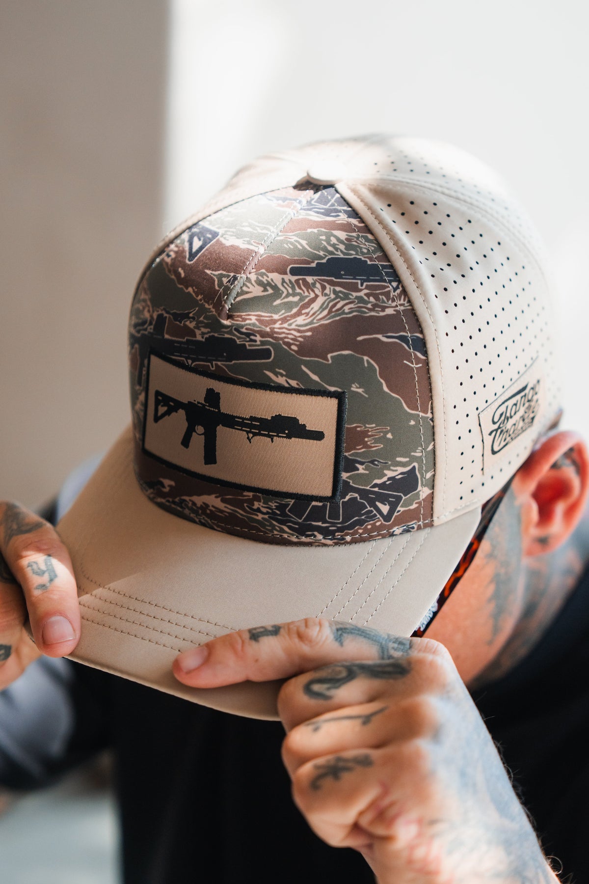 A tattooed person adjusts the Snipes & Stripes Hydro Snapback Hat by Tango Charlie Apparel, featuring a camouflage design, rifle-outline patch, perforated sides, and signature branding.