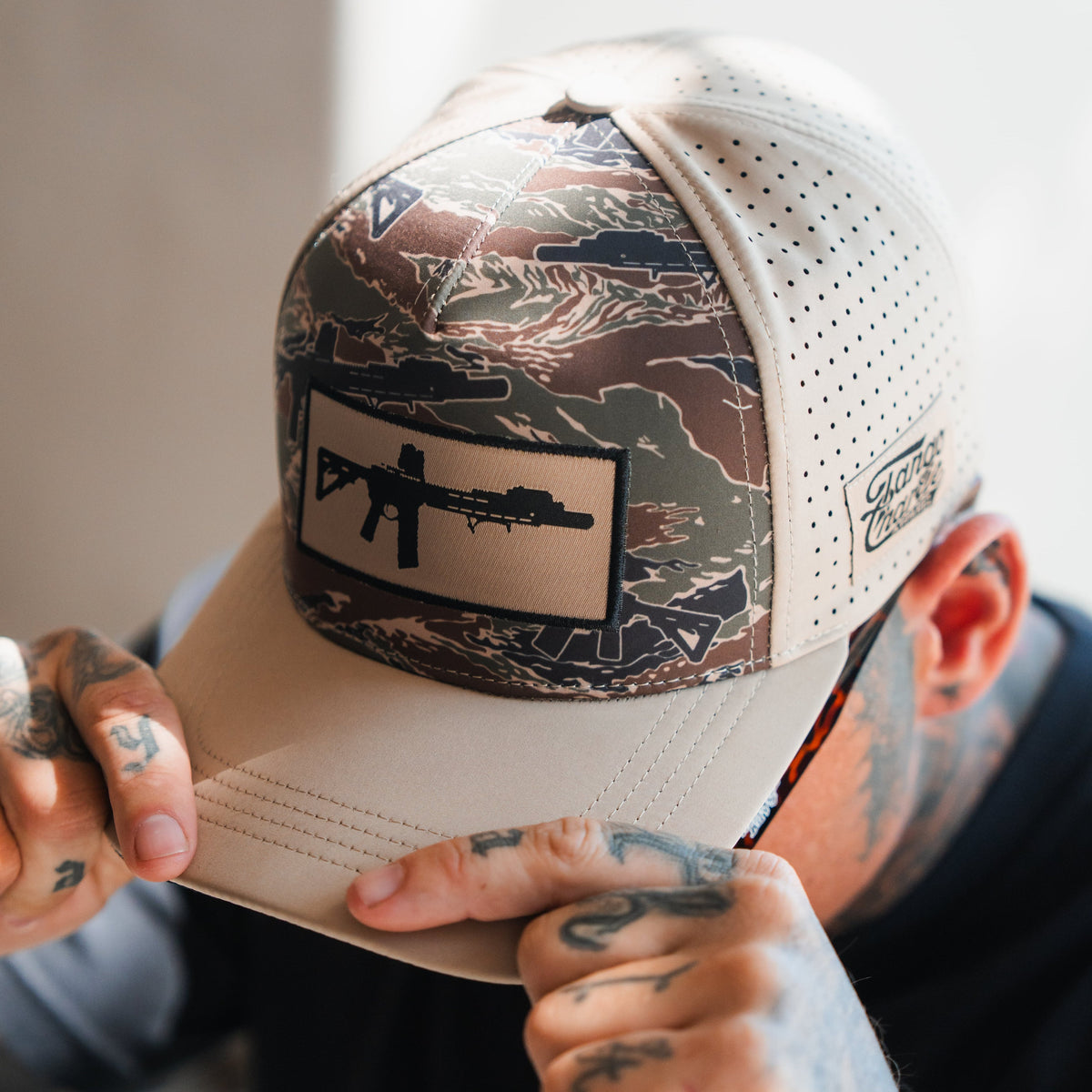 A tattooed individual adjusts a Tango Charlie Apparel Snipes & Stripes Hydro Snapback Hat, featuring camouflage and beige colors with a rifle silhouette patch. Sunlight highlights the cap and hands while their face is mostly obscured.