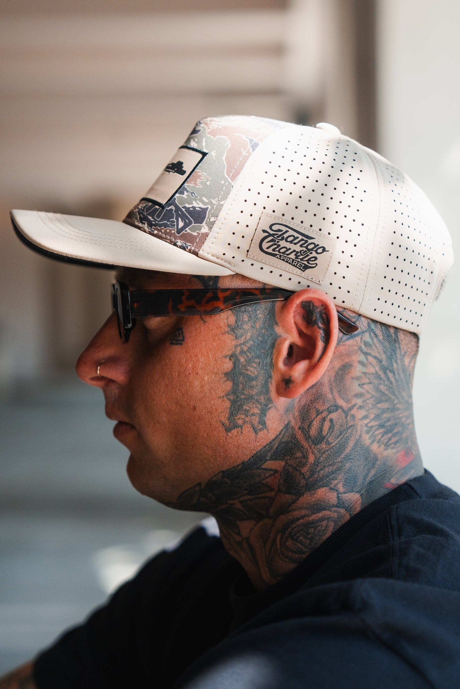 A tattooed person wears the Snipes & Stripes Hydro Snapback Hat by Tango Charlie Apparel, featuring a beige color and patterned brim, along with dark glasses and a nose ring, in front of a blurred background.