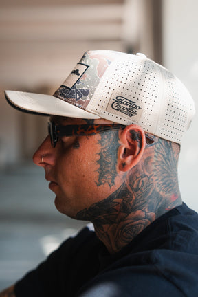 A tattooed person wears the Snipes & Stripes Hydro Snapback Hat by Tango Charlie Apparel, featuring a beige color and patterned brim, along with dark glasses and a nose ring, in front of a blurred background.