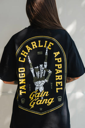 Someone wears the Gain Gang - Premium Oversized Tee by Tango Charlie Apparel, a black oversized shirt with bold yellow lettering and a skeletal hand graphic on the back, featuring standout design and premium details.