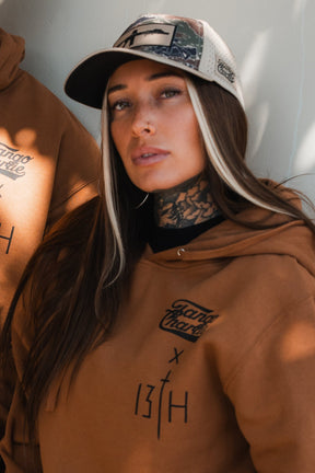 A woman with long dark hair and light streaks, wearing a brown hoodie and the Snipes & Stripes Hydro Snapback Hat by Tango Charlie Apparel, poses against a light background, showing tattoos on her neck.