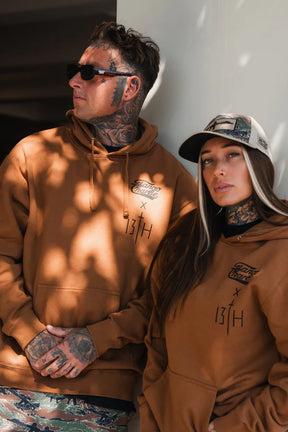 Two people with visible tattoos wear brown "Lift Heavy. Pray Hard." hoodies by Tango Charlie Apparel and casual hats. The person on the right, in a Tango Charlie cap, looks at the camera; the other, in sunglasses, looks away. Sunlight filters over them.