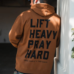 Someone wearing the Lift Heavy. Pray Hard. - Hoodie by Tango Charlie Apparel, featuring bold black "Lift Heavy Pray Hard" text and branding on the back, stands outside leaning against a white wall.