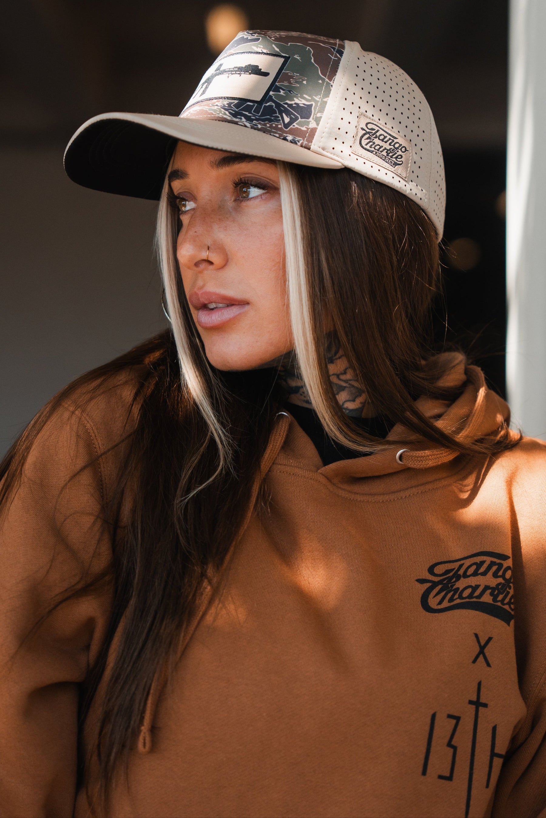 A woman with long brown hair, a nose ring, and neck tattoos wears a tan hoodie and the Snipes & Stripes Hydro - Snapback Hat by Tango Charlie Apparel, gazing thoughtfully to the side in soft indoor light.