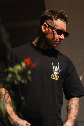 A tattooed man in sunglasses wears the Gain Gang - Premium Oversized Tee by Tango Charlie Apparel—a heavyweight unisex cotton t-shirt with a white hand symbol and yellow text—standing outdoors with blurred red flowers in the foreground.