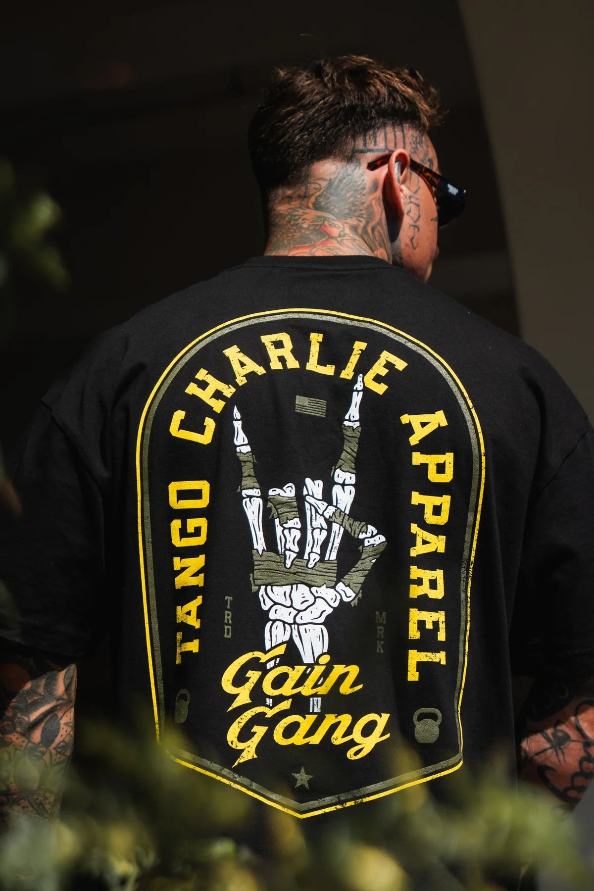 A tattooed individual with short hair and sunglasses models the Tango Charlie Apparel Gain Gang - Premium Oversized Tee, a black unisex heavyweight t-shirt with a bold yellow-outlined skeleton hand and prominent Gain Gang branding.