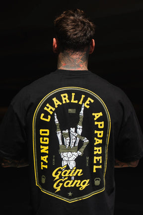 A person with neck tattoos faces away, wearing the Gain Gang - Premium Oversized Tee by Tango Charlie Apparel, featuring a large, colorful back graphic with a hand sign and bold lettering.