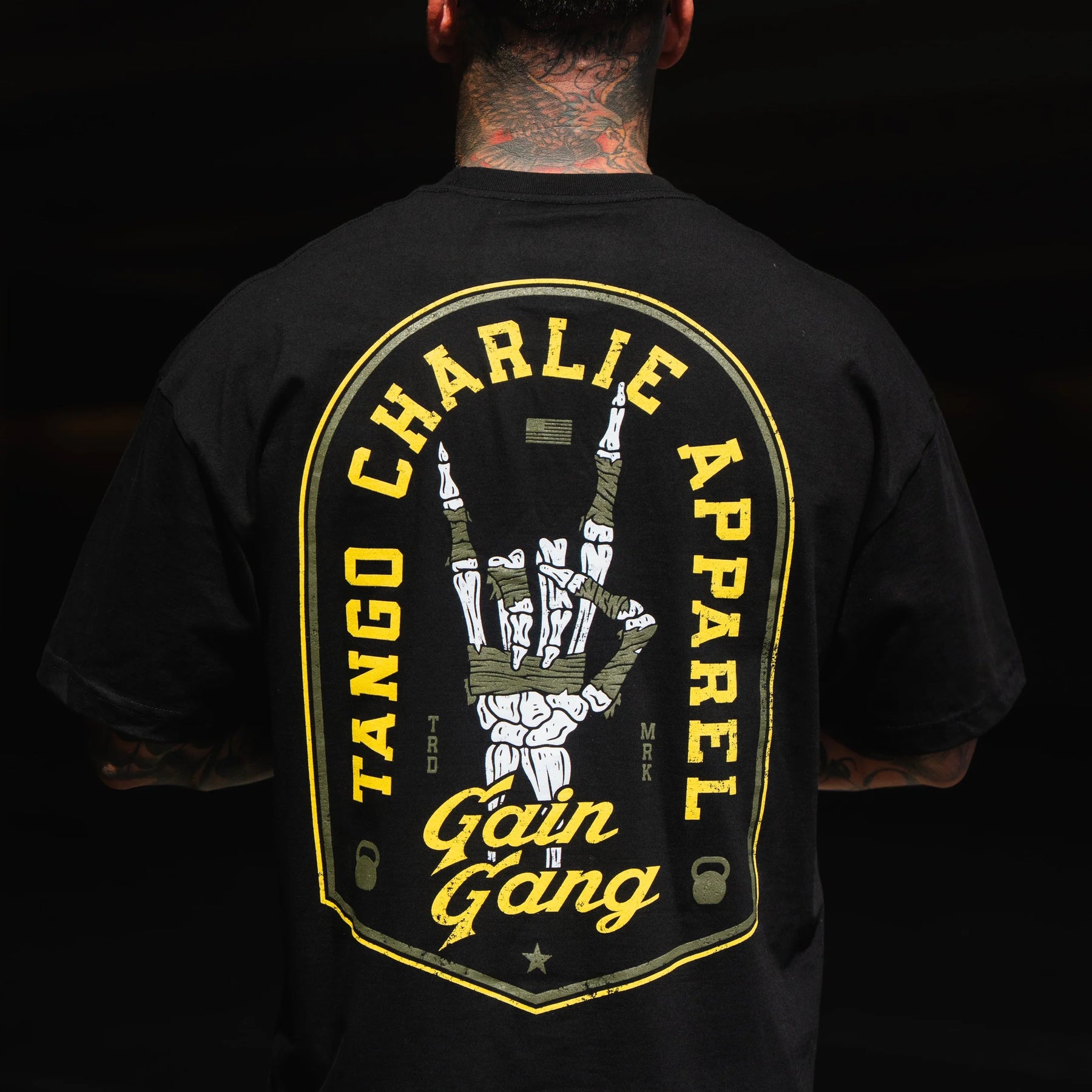 A person wears the Gain Gang - Premium Oversized Tee by Tango Charlie Apparel, featuring a large yellow and white back graphic with a skeleton hand gesture and bold "Gain Gang" branding.