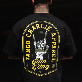 A person wears the Gain Gang - Premium Oversized Tee by Tango Charlie Apparel, featuring a large yellow and white back graphic with a skeleton hand gesture and bold "Gain Gang" branding.