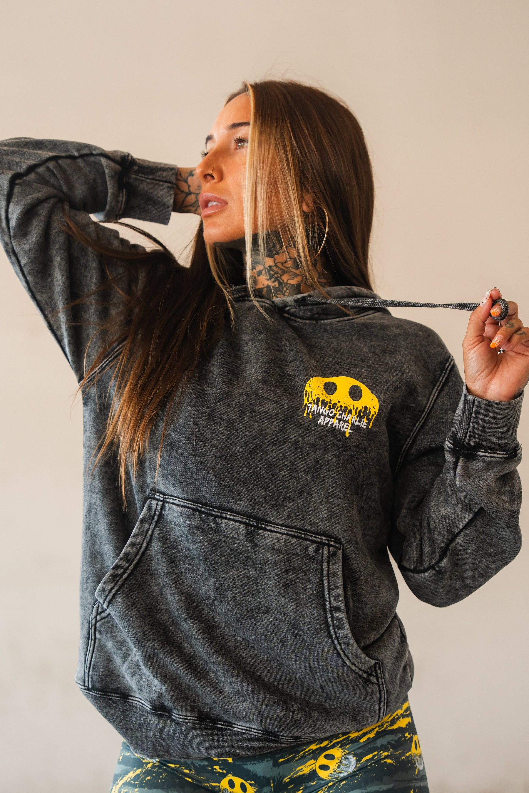 A woman with long brown hair and tattoos poses in the Unleash The Savage Premium Hoodie by Tango Charlie Apparel, featuring a yellow melting smiley face graphic, matching leggings, and holding the hoodie’s drawstring while looking aside.