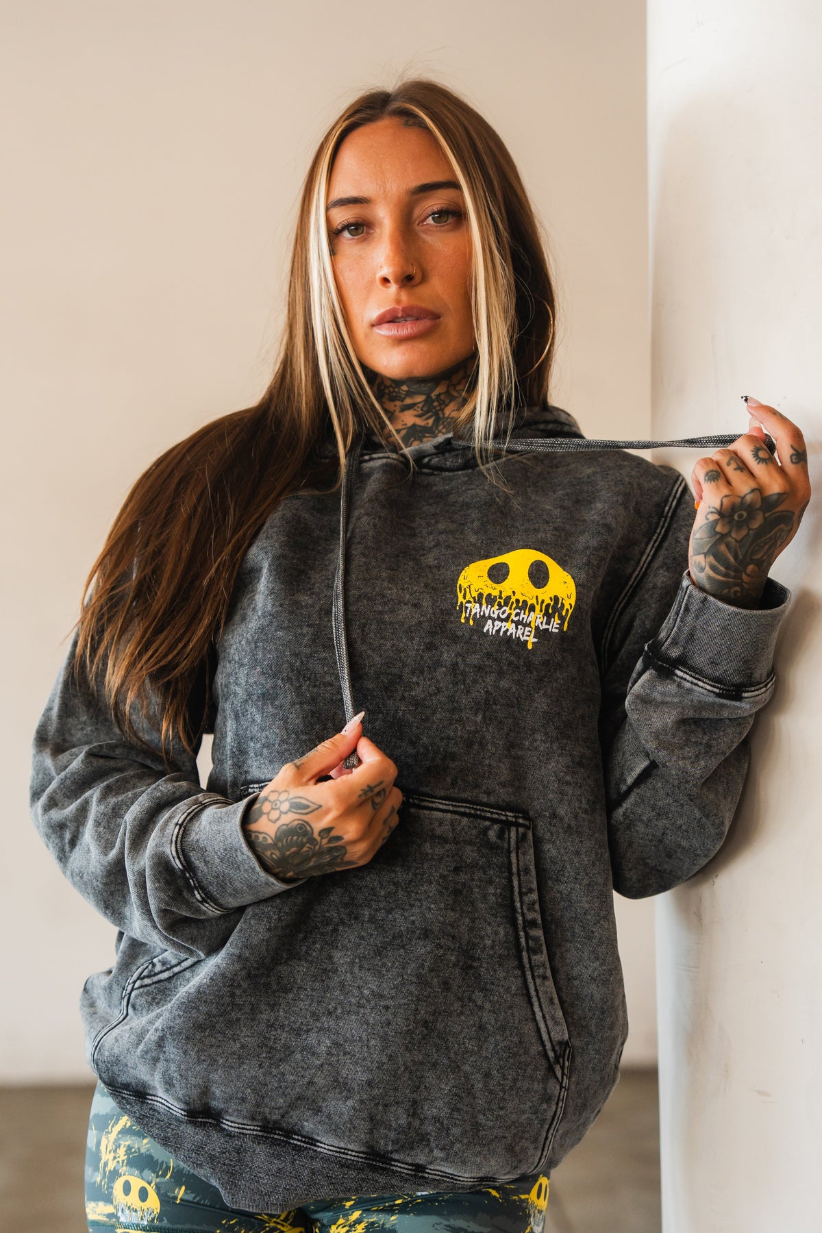 A woman with long brown hair and tattoos wears the Unleash The Savage Premium Hoodie by Tango Charlie Apparel, featuring a yellow graphic, with matching leggings. She stands indoors holding one drawstring and leans against a white wall.