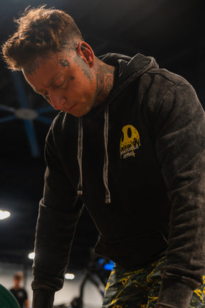 A man with tattoos on his face and neck is looking down in a gym while wearing the Tango Charlie Apparel "Unleash The Savage Premium Hoodie," which features a yellow melting smiley face logo, paired with patterned pants.