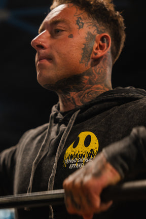 A tattooed individual wears the Unleash The Savage Premium Hoodie by Tango Charlie Apparel, which displays a fierce smile graphic, while gripping a metal bar in a gym setting.