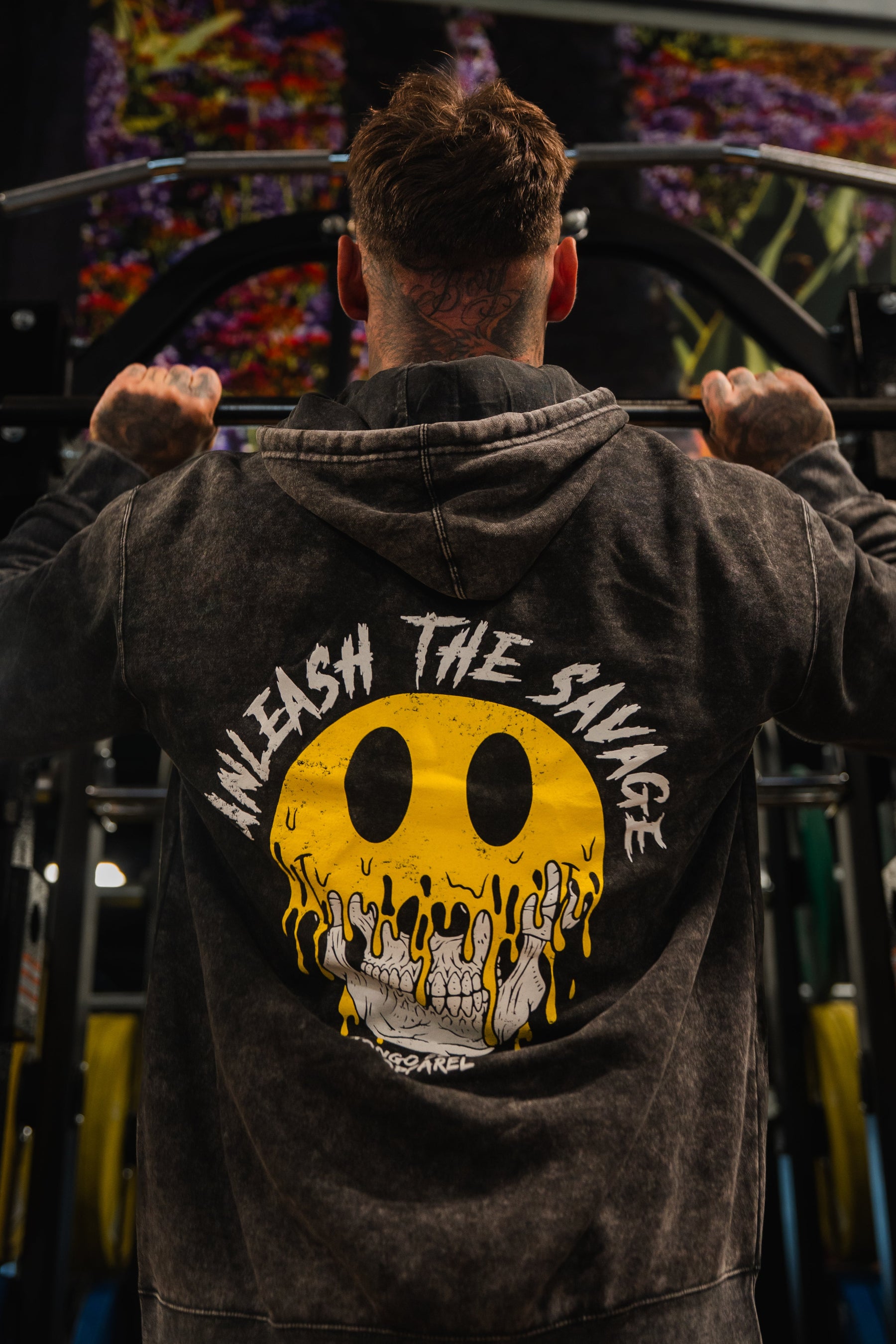 In a gym, someone does pull-ups wearing the Tango Charlie Apparel Unleash The Savage Premium Hoodie, featuring a melting yellow smiley face skull and "UNLEASH THE SAVAGE" printed on the back.