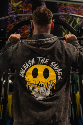 In a gym, someone does pull-ups wearing the Tango Charlie Apparel Unleash The Savage Premium Hoodie, featuring a melting yellow smiley face skull and "UNLEASH THE SAVAGE" printed on the back.