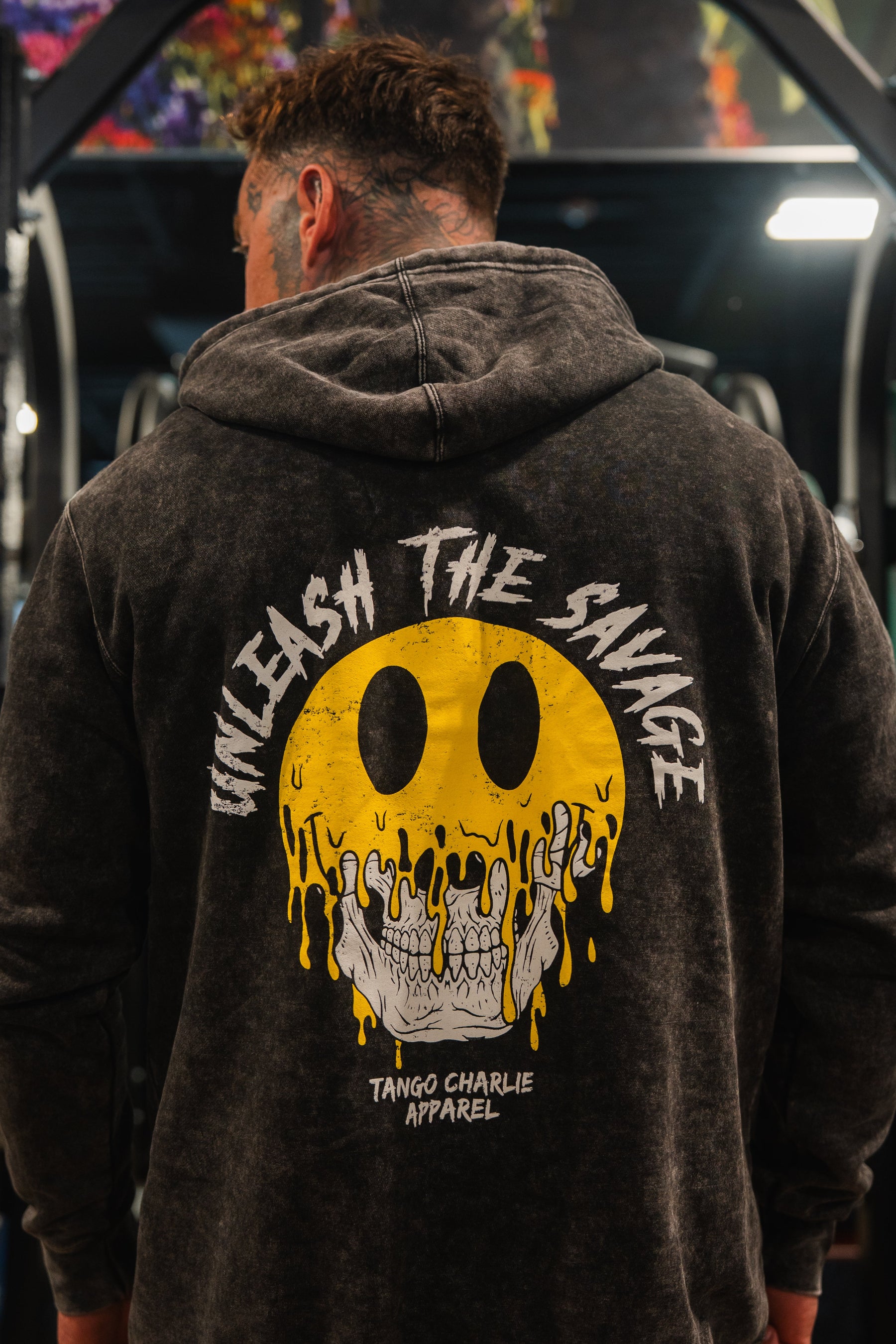 A man at the gym, seen from behind, wearing the Unleash The Savage Premium Hoodie by Tango Charlie Apparel—featuring "Unleash the Savage" above a melting smile skull graphic and the brand name below—ideal for expressing your authentic self.