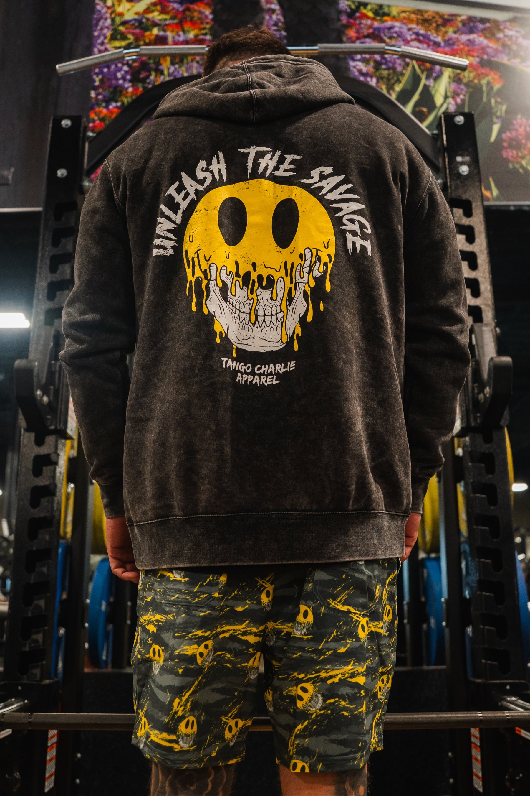 In a gym rack, someone rocks the Tango Charlie Apparel Unleash The Savage Premium Hoodie—featuring a melting smiley and bold back print—paired with yellow patterned shorts for a fearless, authentic look.