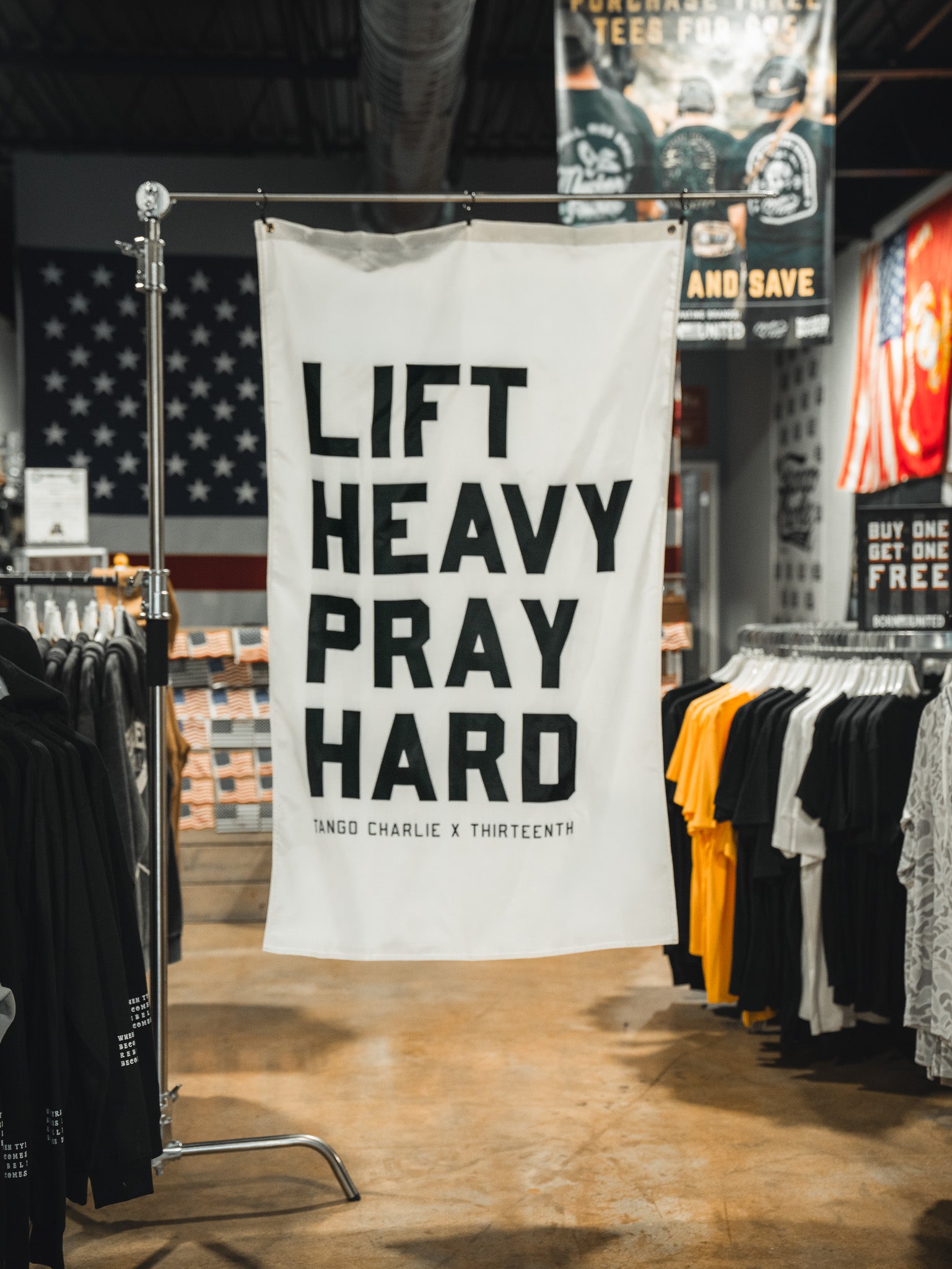 A white Tango Charlie Apparel "Lift Heavy. Pray Hard. Flag" with bold black text hangs in a clothing store, surrounded by shirt racks and an American flag—a striking display that unites faith and fitness.