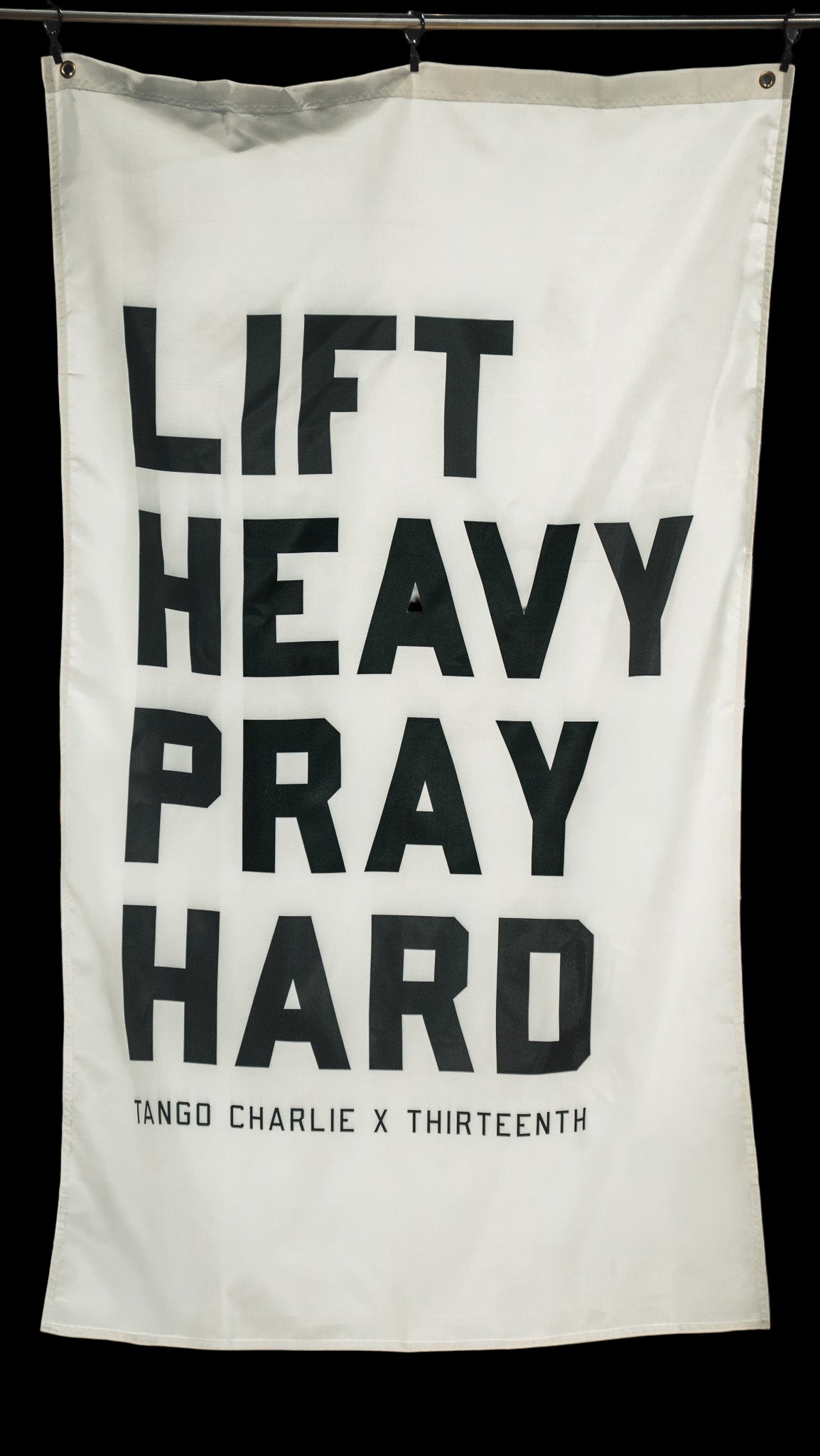 A white "Lift Heavy. Pray Hard." flag by Tango Charlie Apparel features bold black text and hangs in a store with clothing and American flag decor, symbolizing a blend of faith and fitness.