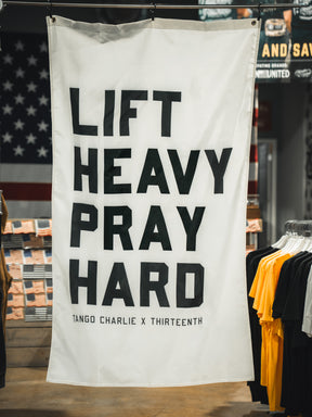 A white "Lift Heavy. Pray Hard." flag by Tango Charlie Apparel features bold black text and hangs in a store with clothing and American flag decor, symbolizing a blend of faith and fitness.