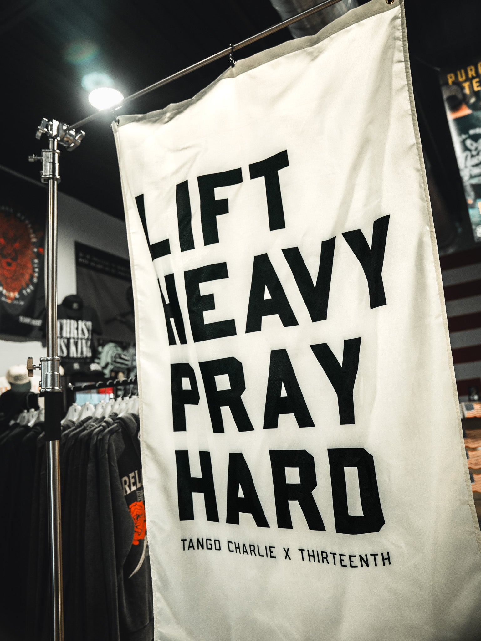 A white Lift Heavy. Pray Hard. Flag by Tango Charlie Apparel with bold black text hangs in a clothing store, surrounded by shirts and a U.S. flag, inspiring faith and fitness.
