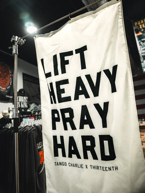 A white Lift Heavy. Pray Hard. Flag by Tango Charlie Apparel with bold black text hangs in a clothing store, surrounded by shirts and a U.S. flag, inspiring faith and fitness.