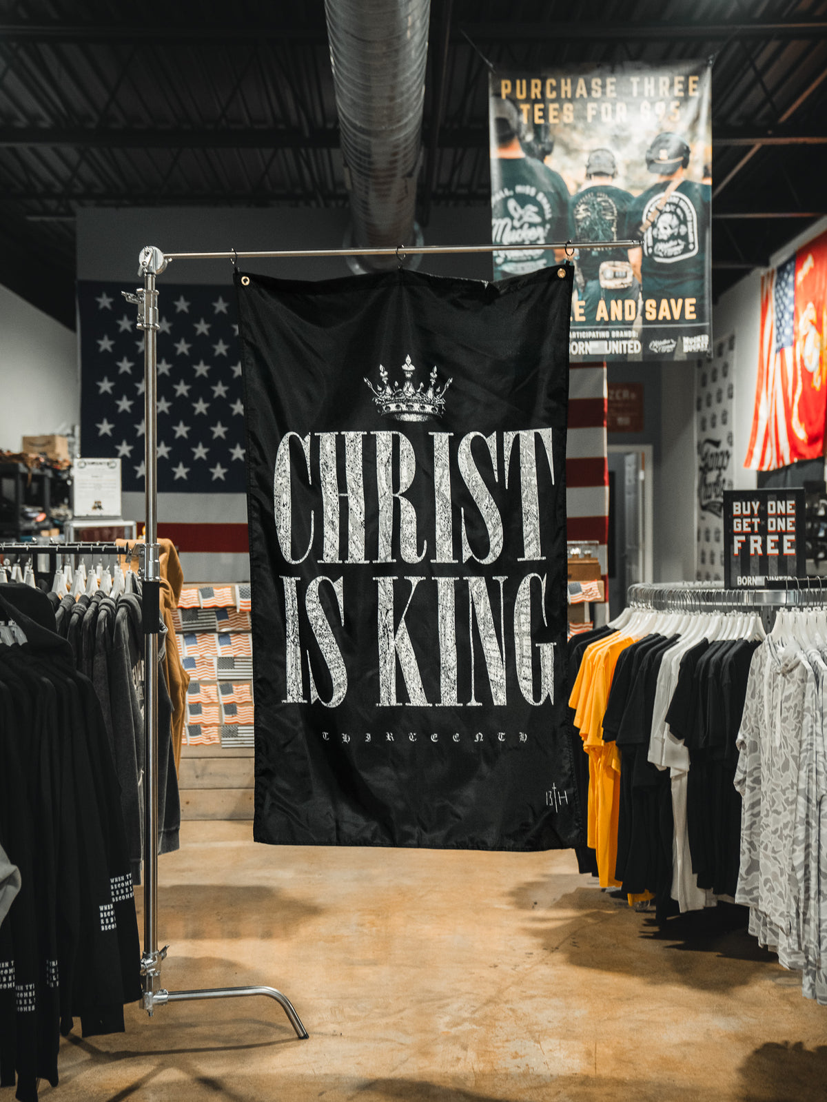A black 13th Christ is King Flag with bold white letters hangs in a clothing store, displayed among t-shirts and a large American flag on the back wall.