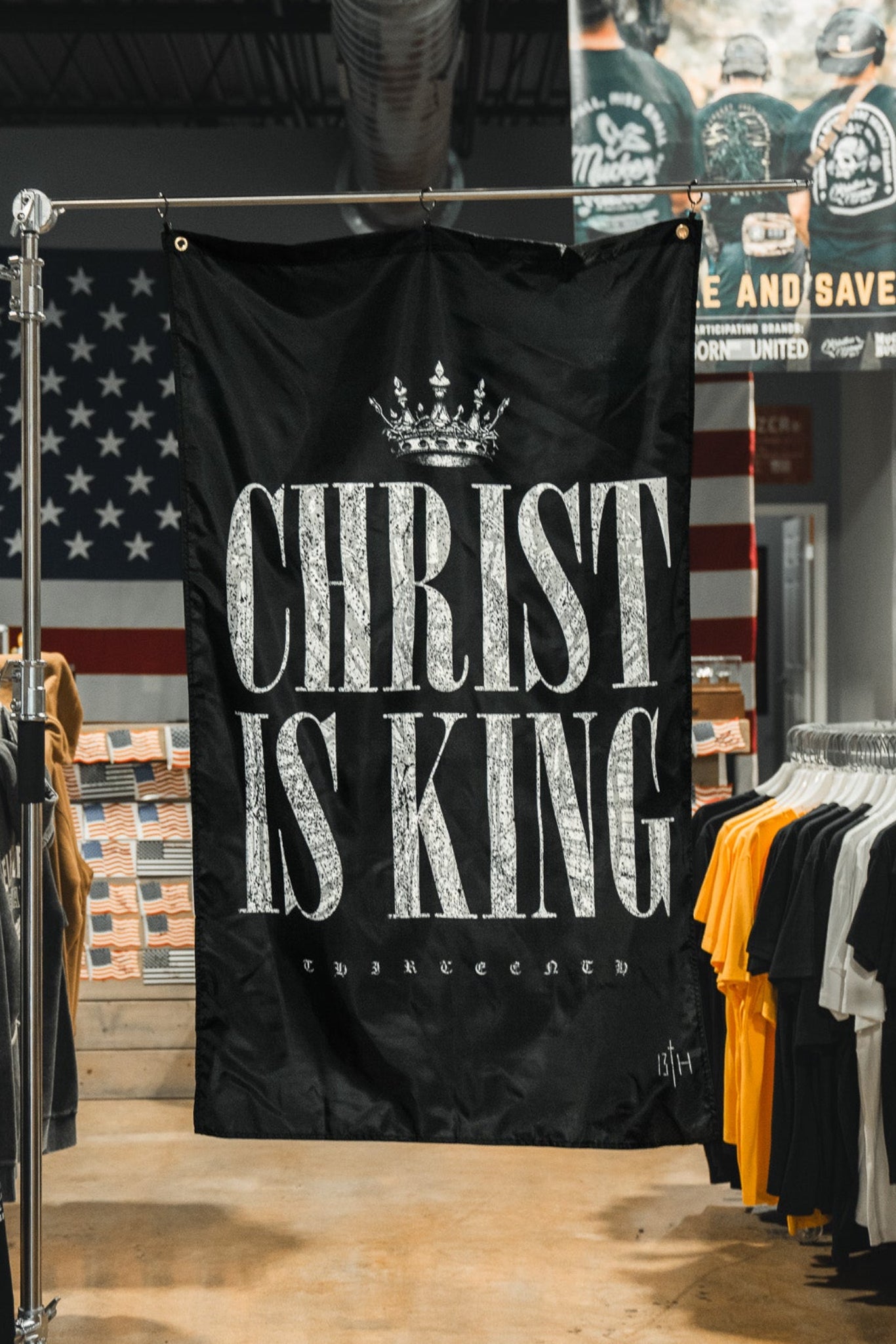 A black 13th Christ is King Flag with bold white letters hangs in a clothing store, displayed among t-shirts and a large American flag on the back wall.