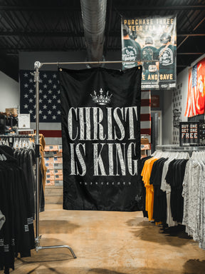 A black 13th Christ is King Flag with bold white letters hangs in a clothing store, displayed among t-shirts and a large American flag on the back wall.