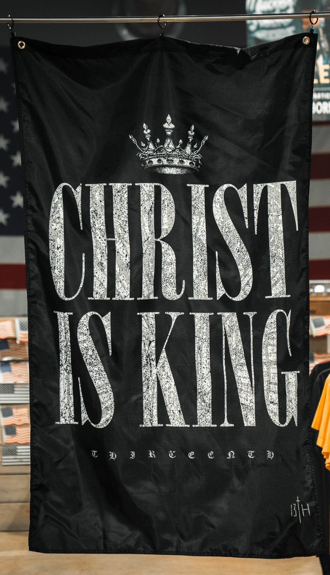 A 13th Christ is King Flag, featuring black fabric with white lettering and a crown, is showcased in a clothing store amid shirt racks and an American flag in the background.