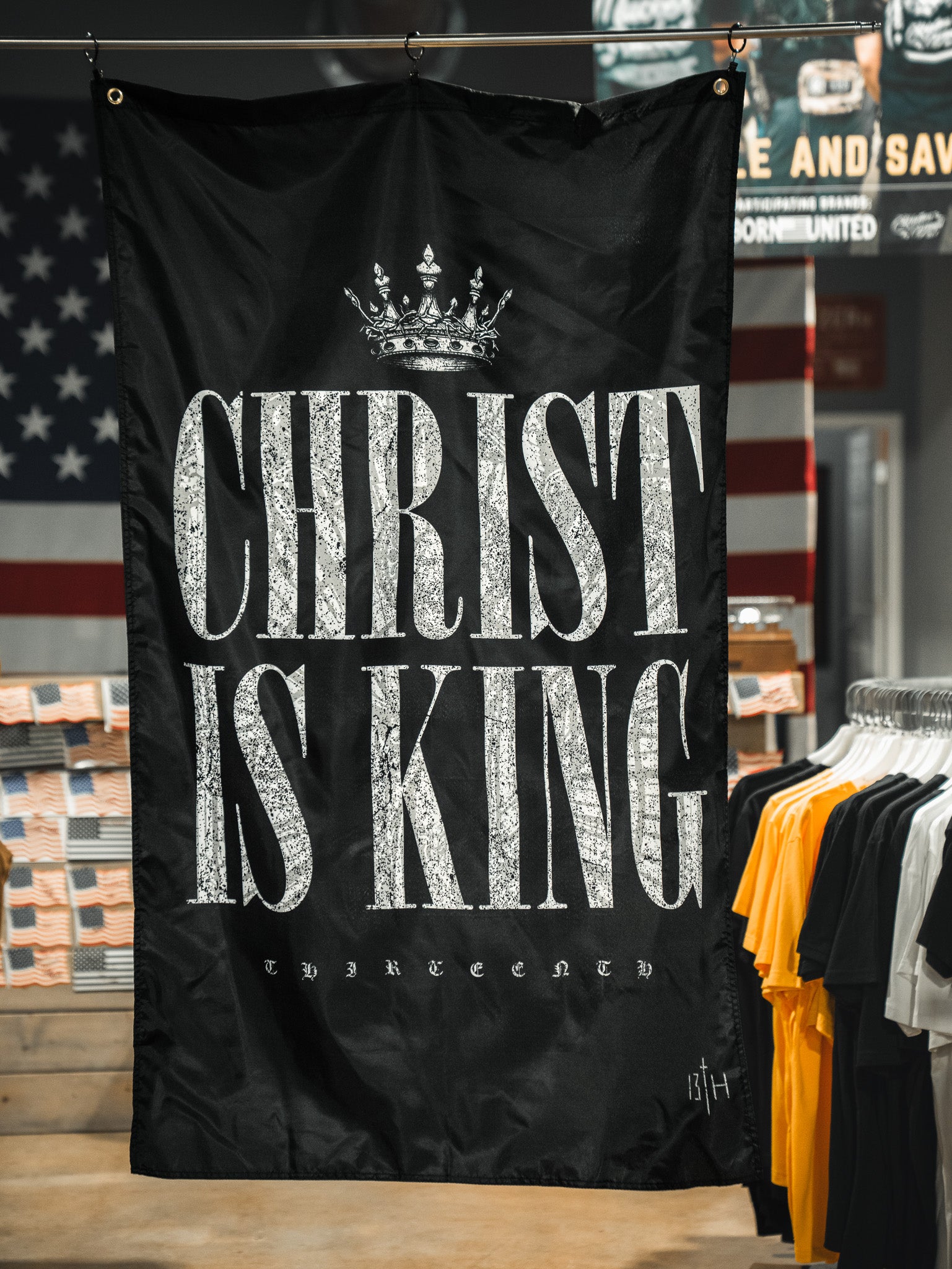 A 13th Christ is King Flag, featuring black fabric with white lettering and a crown, is showcased in a clothing store amid shirt racks and an American flag in the background.