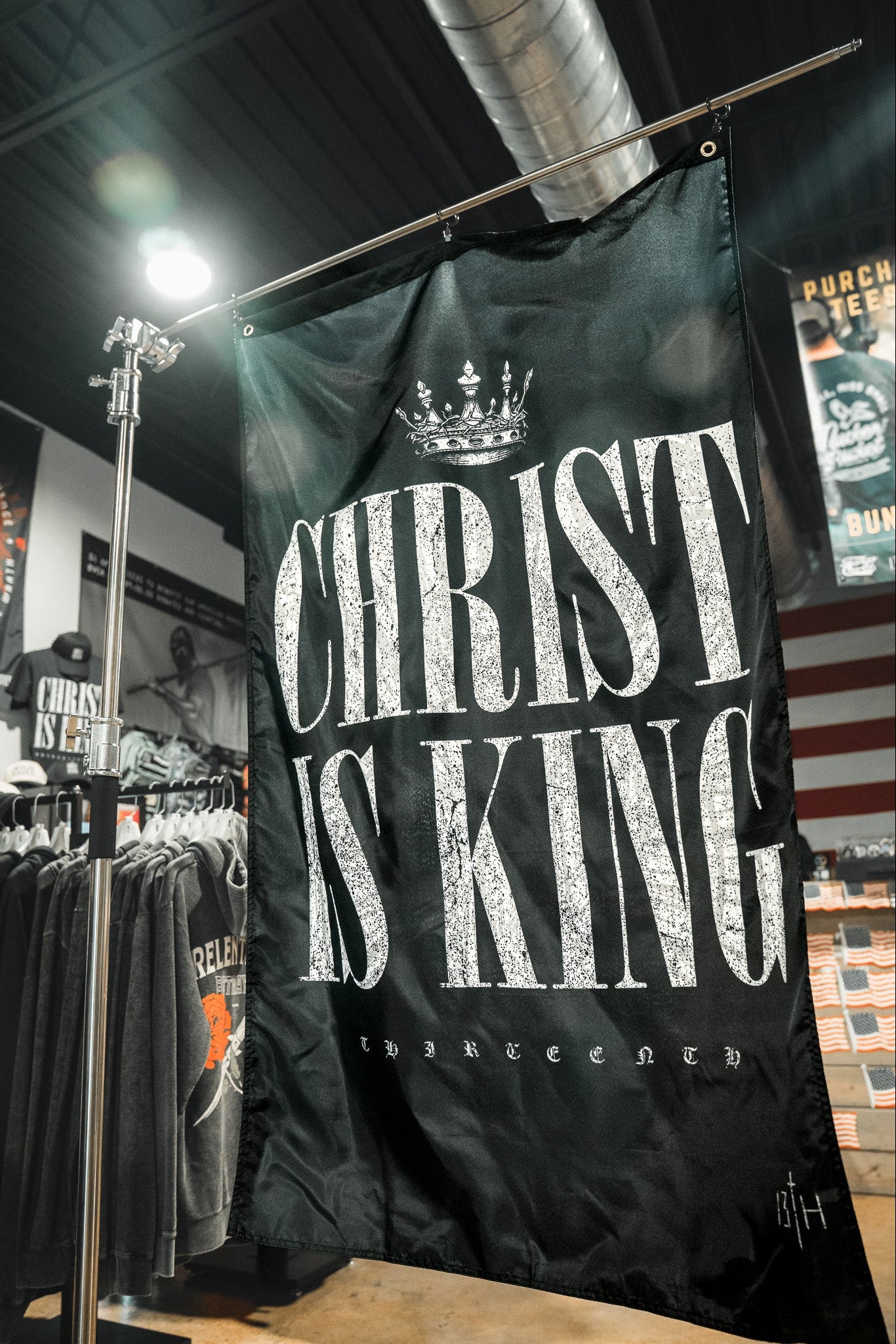 The 13th Christ is King Flag, a large black banner with a crown graphic and bold white text, hangs in a clothing store surrounded by racks of shirts and other merchandise.