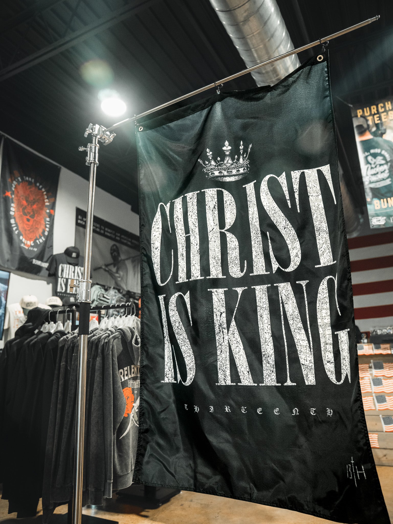 The 13th Christ is King Flag, a large black banner with a crown graphic and bold white text, hangs in a clothing store surrounded by racks of shirts and other merchandise.
