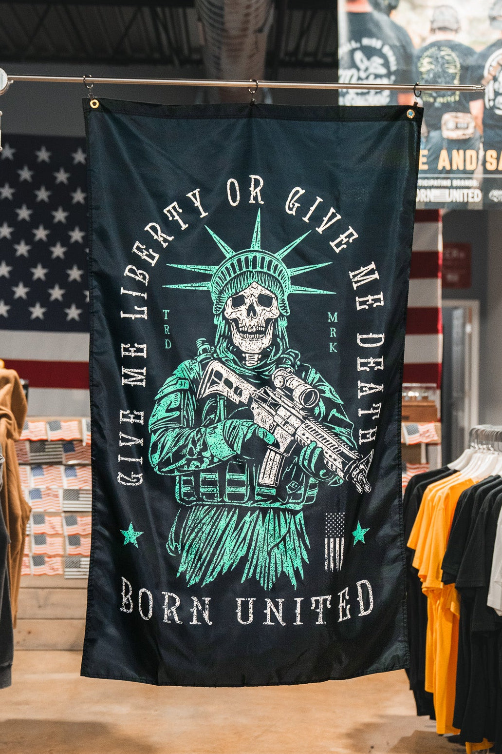 A Liberty or Death Flag by Born United Private Label, showing a skeleton in a Statue of Liberty crown with a rifle and the text “Give Me Liberty Or Give Me Death, Born United,” hangs in a clothing store with American flags and T-shirts in the background.