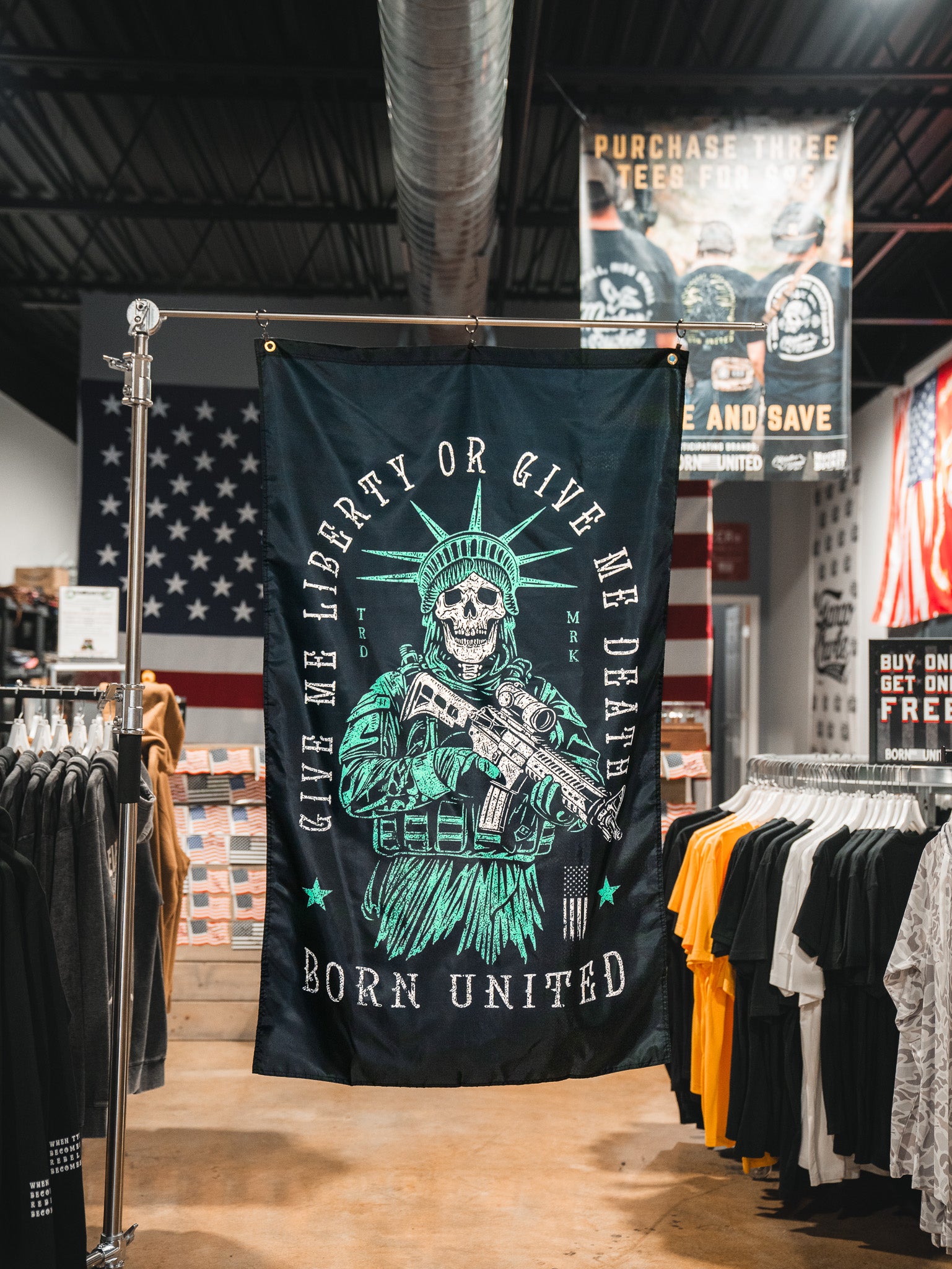 A Liberty or Death Flag by Born United Private Label, showing a skeleton in a Statue of Liberty crown with a rifle and the text “Give Me Liberty Or Give Me Death, Born United,” hangs in a clothing store with American flags and T-shirts in the background.
