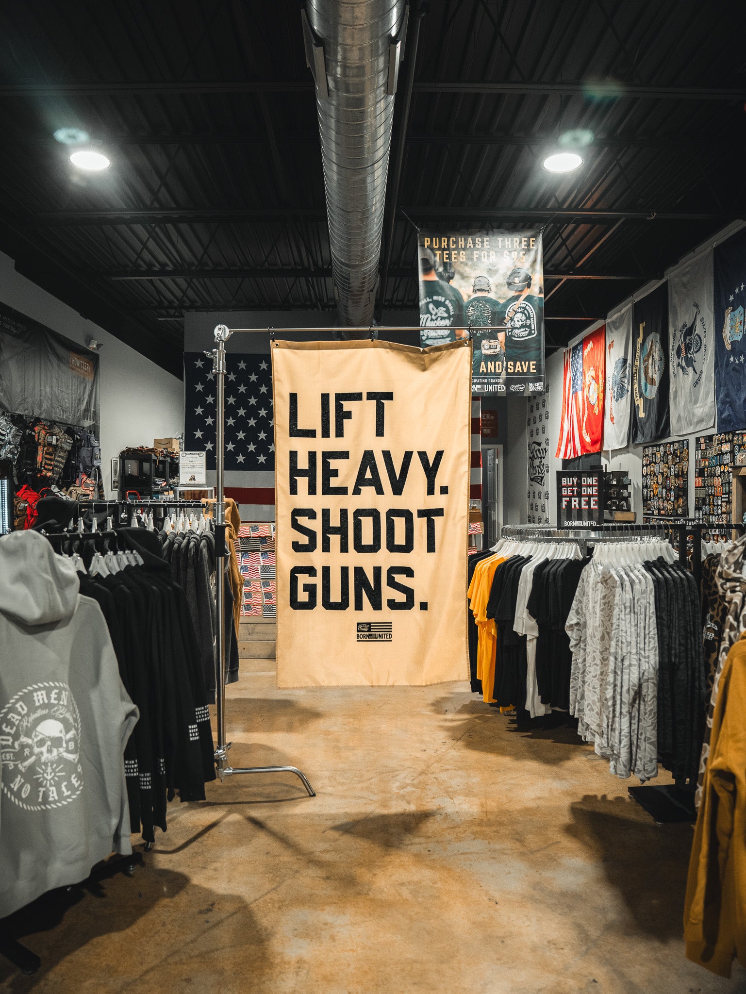 A large beige "Lift Heavy. Shoot Guns." flag by Tango Charlie Apparel hangs in a store stocked with strength training shirts, hoodies, posters, and patriotic flags.