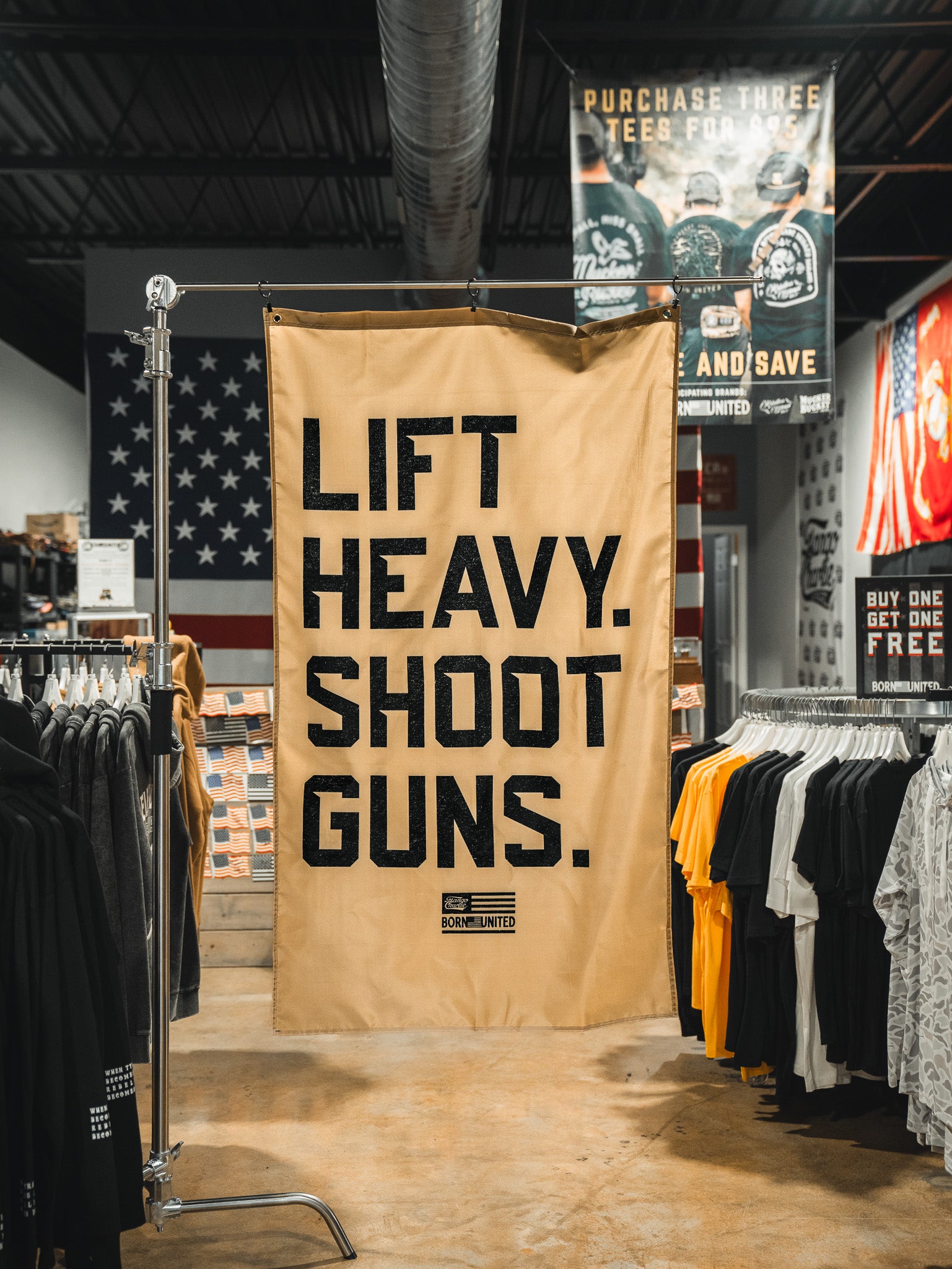 A Lift Heavy. Shoot Guns. Flag by Tango Charlie Apparel hangs in a clothing store above t-shirt racks, with bold black lettering and a patriotic flag on the wall in the background.