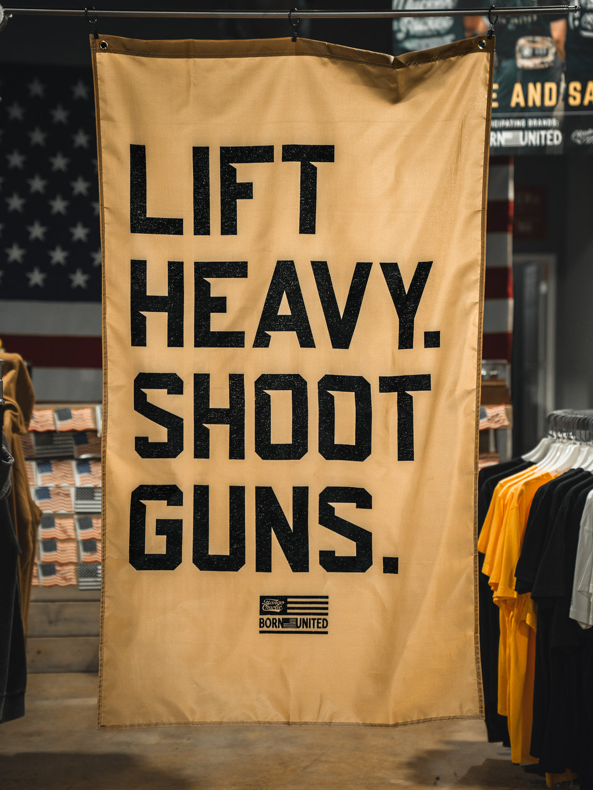 A tan "Lift Heavy. Shoot Guns. Flag" by Tango Charlie Apparel hangs indoors among clothing racks, featuring bold black text and a patriotic flag design that embodies strength training and shooting sports.
