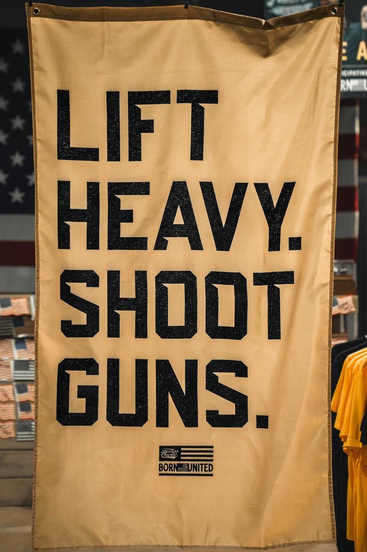 A tan "Lift Heavy. Shoot Guns. Flag" by Tango Charlie Apparel hangs indoors among clothing racks, featuring bold black text and a patriotic flag design that embodies strength training and shooting sports.