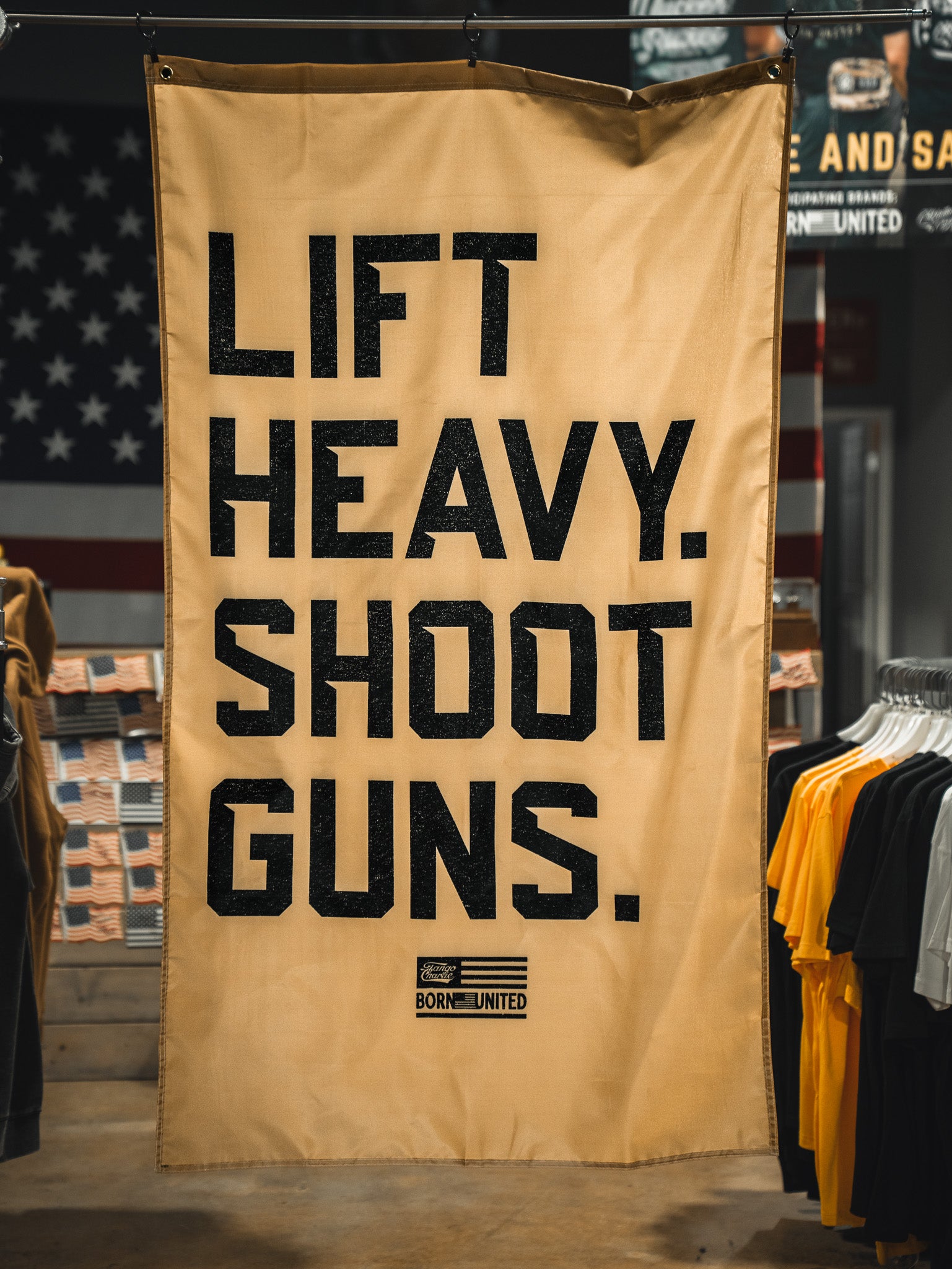 A tan "Lift Heavy. Shoot Guns. Flag" by Tango Charlie Apparel hangs indoors among clothing racks, featuring bold black text and a patriotic flag design that embodies strength training and shooting sports.