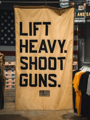 A tan "Lift Heavy. Shoot Guns. Flag" by Tango Charlie Apparel hangs indoors among clothing racks, featuring bold black text and a patriotic flag design that embodies strength training and shooting sports.