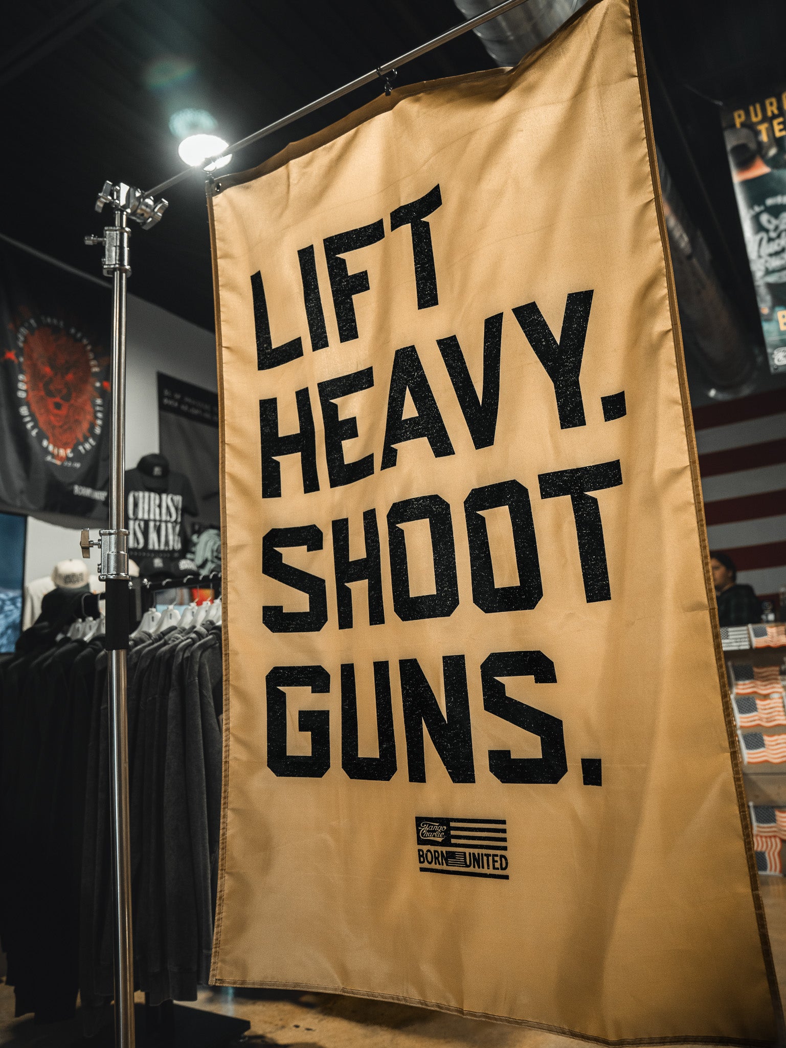 A large tan banner by Tango Charlie Apparel displays bold black text, "LIFT HEAVY. SHOOT GUNS.", with an American flag patch and "BORN UNITED" at the bottom. The product is the Lift Heavy. Shoot Guns. Flag; clothing racks are visible in the background.