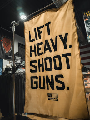 A large tan banner by Tango Charlie Apparel displays bold black text, "LIFT HEAVY. SHOOT GUNS.", with an American flag patch and "BORN UNITED" at the bottom. The product is the Lift Heavy. Shoot Guns. Flag; clothing racks are visible in the background.
