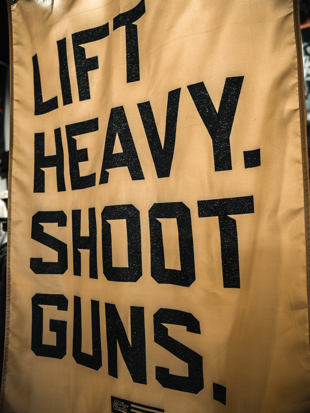 The Lift Heavy. Shoot Guns. Flag by Tango Charlie Apparel features bold black lettering on tan fabric, showcasing your passion for strength training and shooting sports in a striking, eye-catching design.